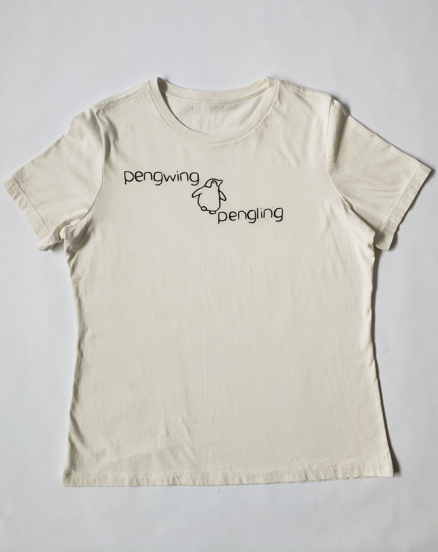 A full shot of the crew neck cotton tee with "pengwing", "pengling", and a cartoonish penguin hand stitched in black thread. The shirt is laid flat on a white background. The font is similar to Helvetica. "pengwing" is stitched to the upper left, "pengling" is stitched to the lower right. The actual penguin is in the middle and looks to plan to walk over "pengling".