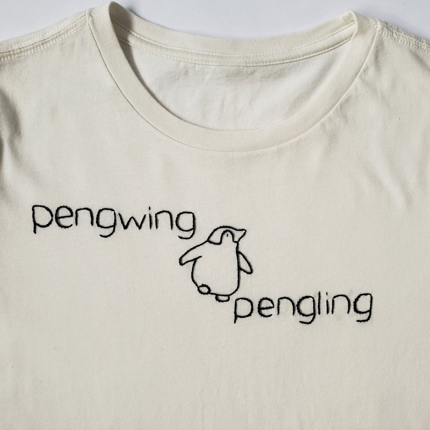 A detail shot of the hand stitching on the soft white cotton shirt. Pengwing is stitched to the upper left, a plump penguin is mid waddle in the middle - his right foot is poised midair. Pengling is stitched on the right of the waddling penguin.