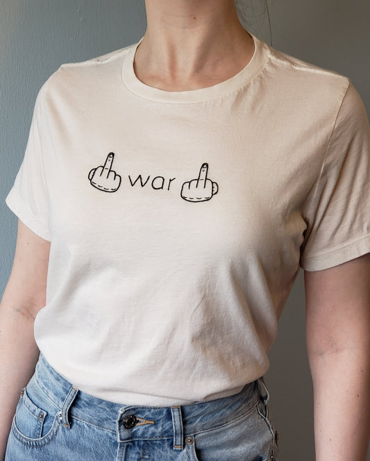 A woman wears a soft white tee tucked into light wash jeans. The shirt has war hand embroidered in black between two hands giving the finger. The hands on the shirt have the thumbs out, as your hands would be if you were flipping war off.