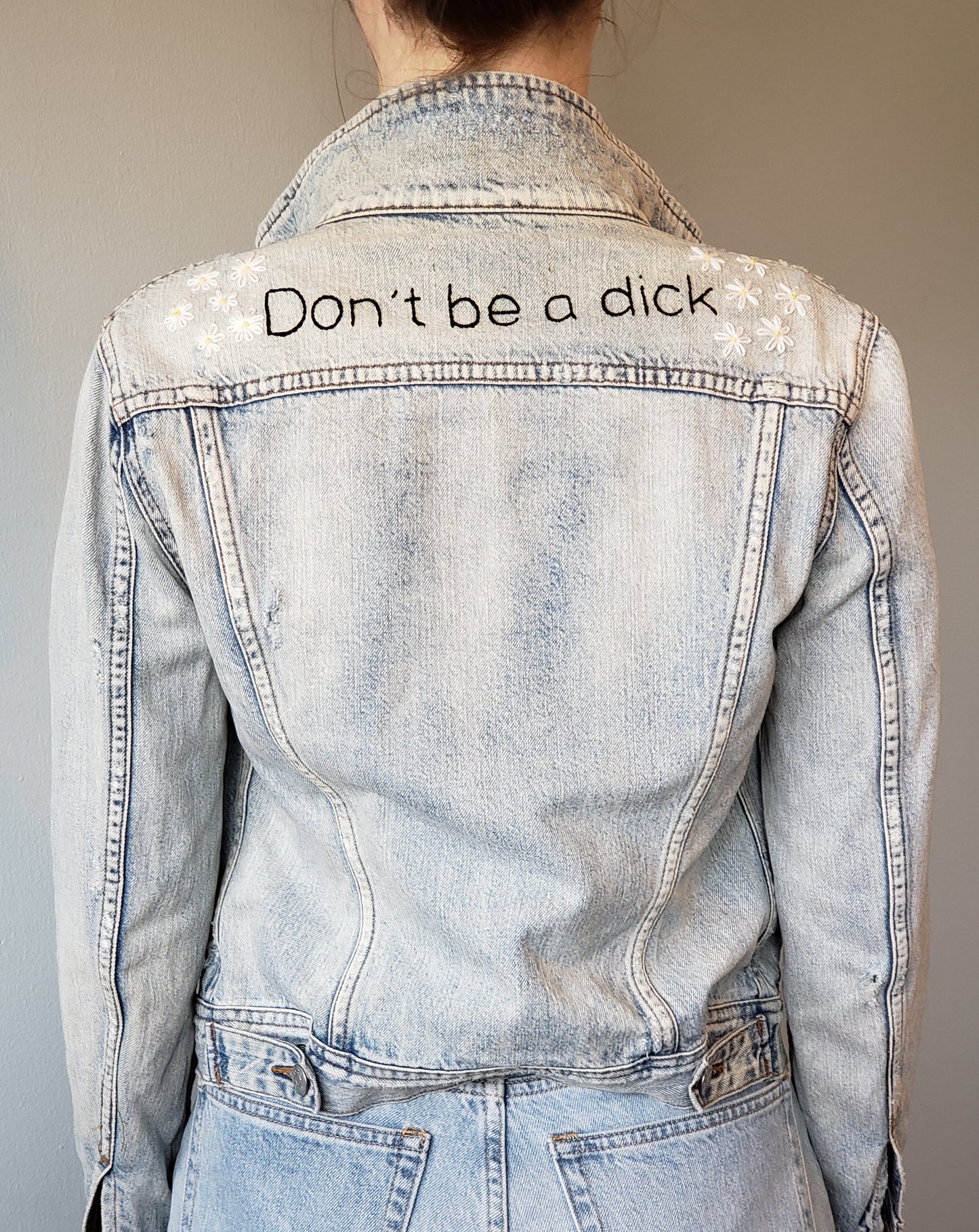 A woman wears an acid wash denim jacket in front of a light grey background. The jacket has "Don't be a dick" hand embroidered in black, and daises sewn on both shoulders. There are 6 daisies on each shoulder.