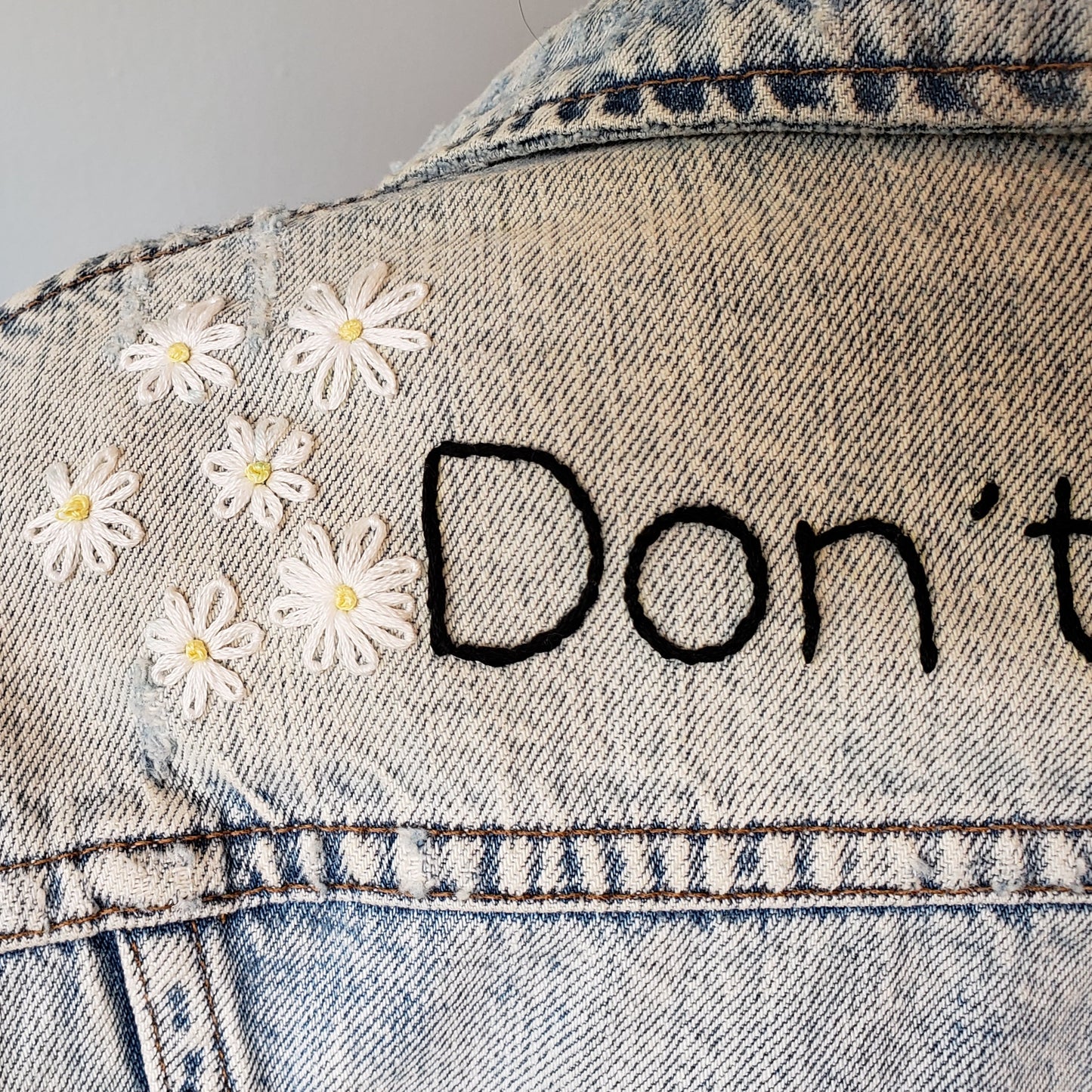 A detail shot of the daisies on the acid wash jacket. We can see the word "Don't" but this shot is about the 6 flowers that sit beside the text. Each flower has a light yellow french knot in the middle, and white petals. Most flowers have 8 petals.