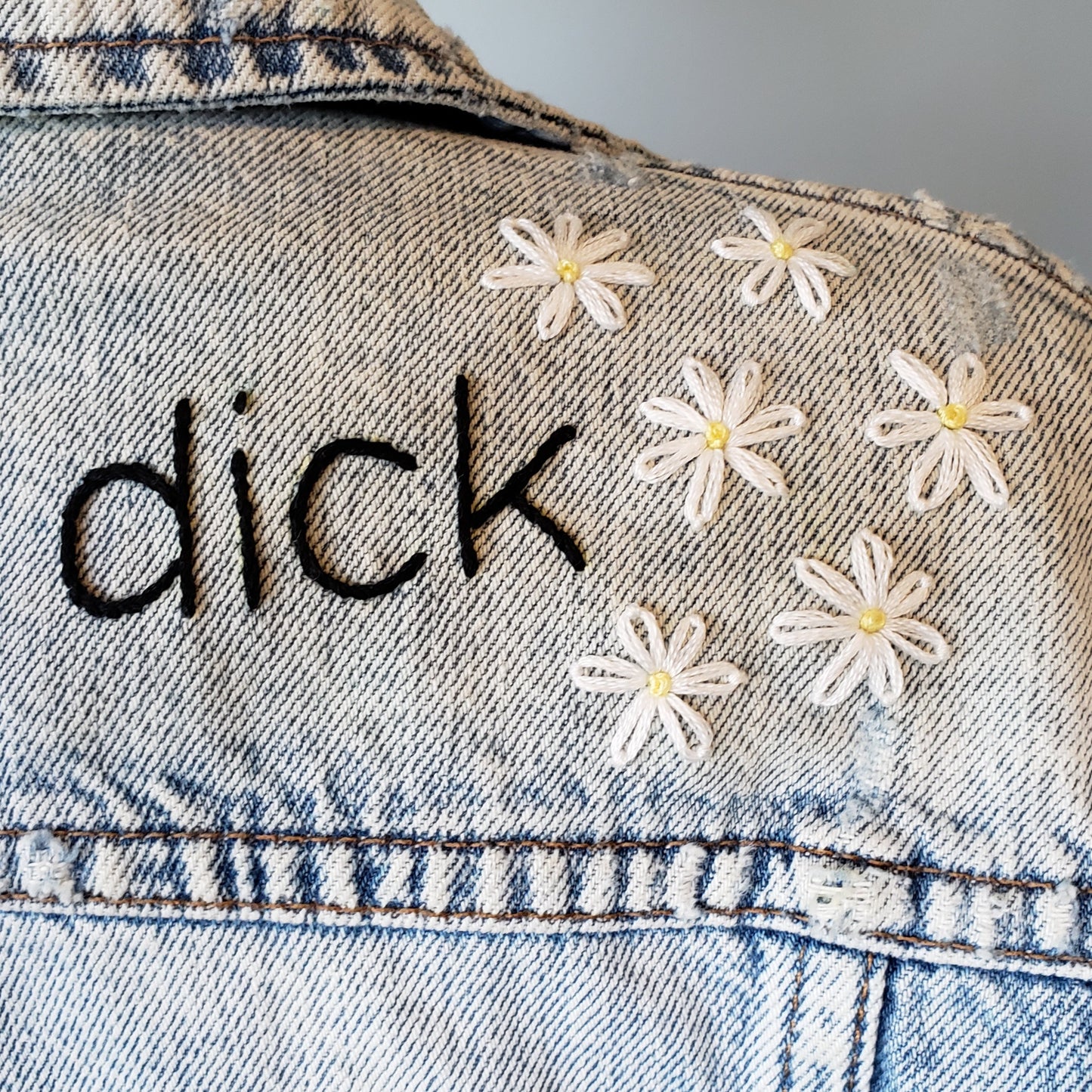 A detail shot of the daisies on the acid wash jacket. We can see the word "dick" but this shot is about the 6 flowers that sit beside the text. Each flower has a yellow french knot in the middle, and white petals. Most flowers have 7 petals.