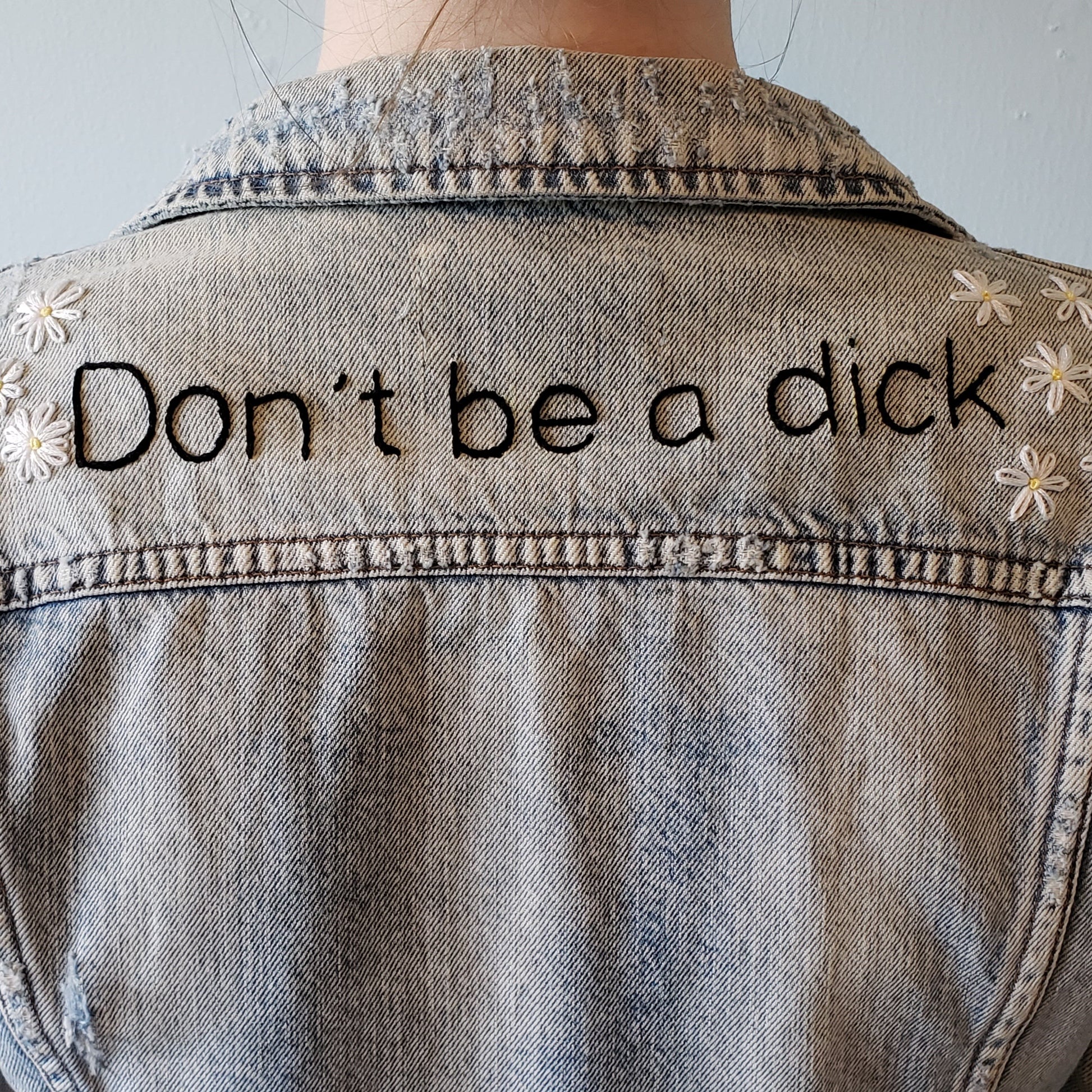 The back of the jacket, this time closer up. "Don't be a dick" is hand sewn straight across the back panel, and 6 daisies bookend the text on each side. The text is stitched in black, and looks quite bold in a font similar to Helvetica.