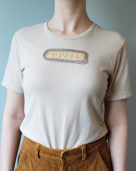 A shot of a woman, taken from the neck to the hip. She wears a light sandstone tee. The crewneck tee has "GOLDEN" hand stitched in a groovy font, with burnished gold thread. Golden is outlined in orange, brown, and green concentric lines.