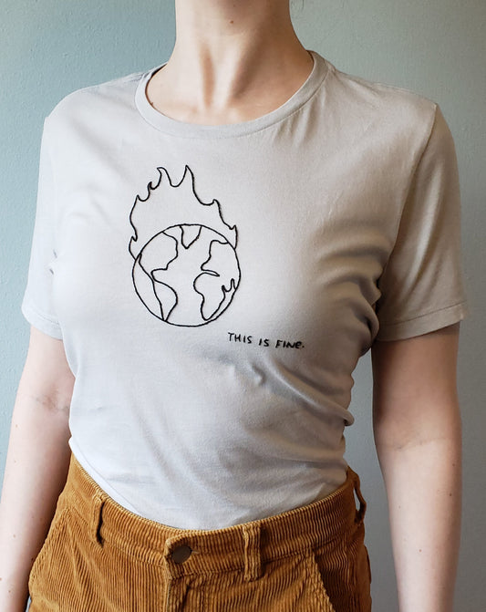 A woman wears a light grey crewneck tee tucked into brown corduroy pants. The shirt has the earth with flames leaping off it stitched in black in the middle of the chest, "this is fine" is stitched on the bottom, to the right, quite small.