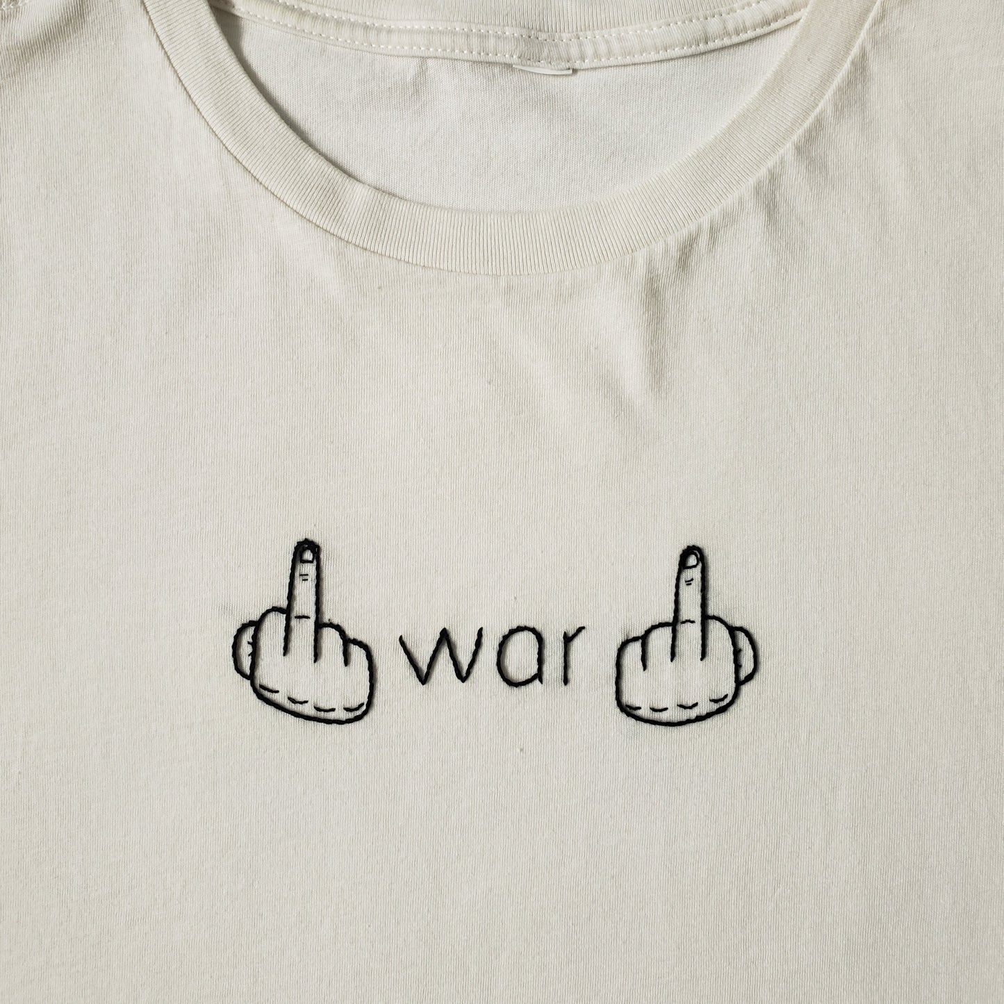 A close up of an off white crew neck tee with war and two hands flipping the bird hand embroidered in black thread. It is all done in a vine stitch. The text is in a font similar to Helvetica and the hands are slightly brutalist in style. We can see that the stitching is even and the hands have detail on the knuckles. 