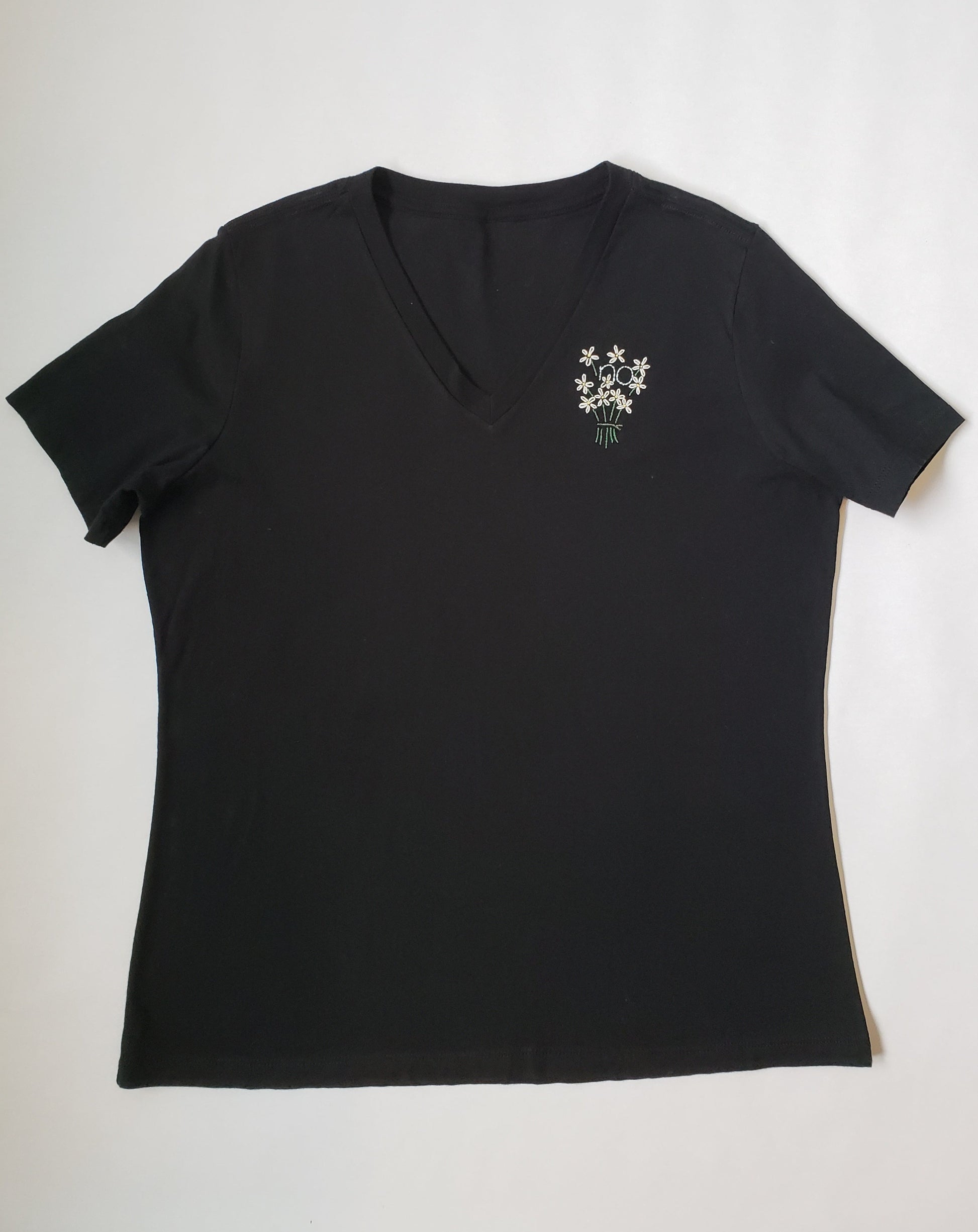A black v neck tee with a small bouquet of hand embroidered flowers on the left side. The bouquet has 10 daisies and "no" embroidered in light blue in a backstitch in the middle of the bouquet. The flowers are held together by a small grey tie.
