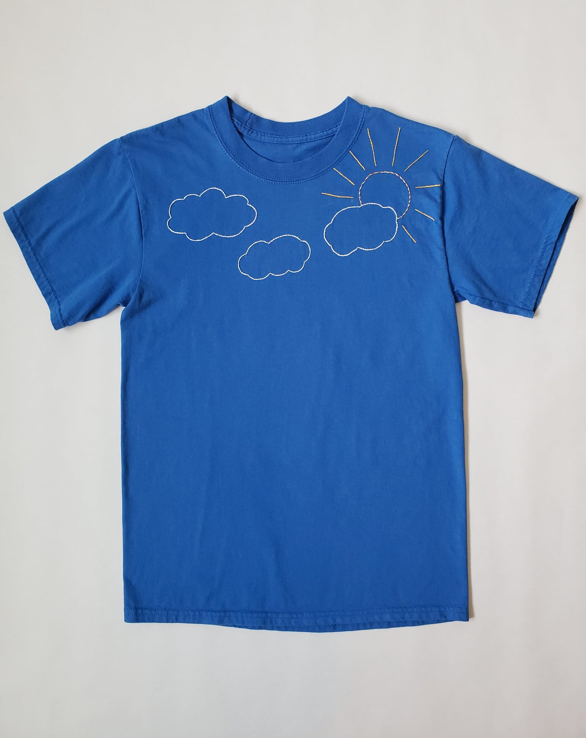 A blue crewneck tee laid flat.  There are three white clouds and a sun stitched in yellow, red, and orange in a twisted effect, with yellow rays. The sun pokes out from behind the rightmost cloud.