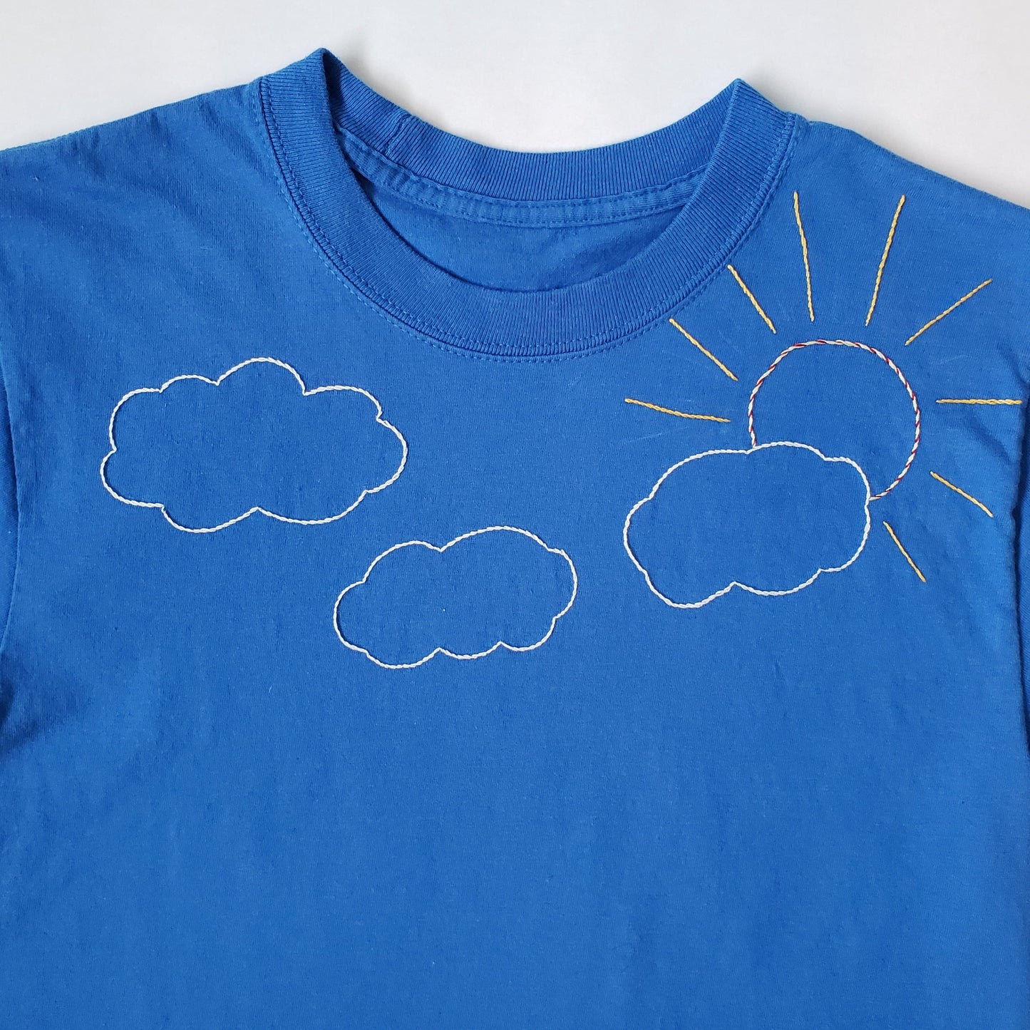 A close up of the embroidery on the chest of the front of the blue crewneck tee. There are three white clouds and a sun stitched in yellow, red, and orange in a twisted effect, with yellow rays. The sun pokes out from behind the rightmost cloud.