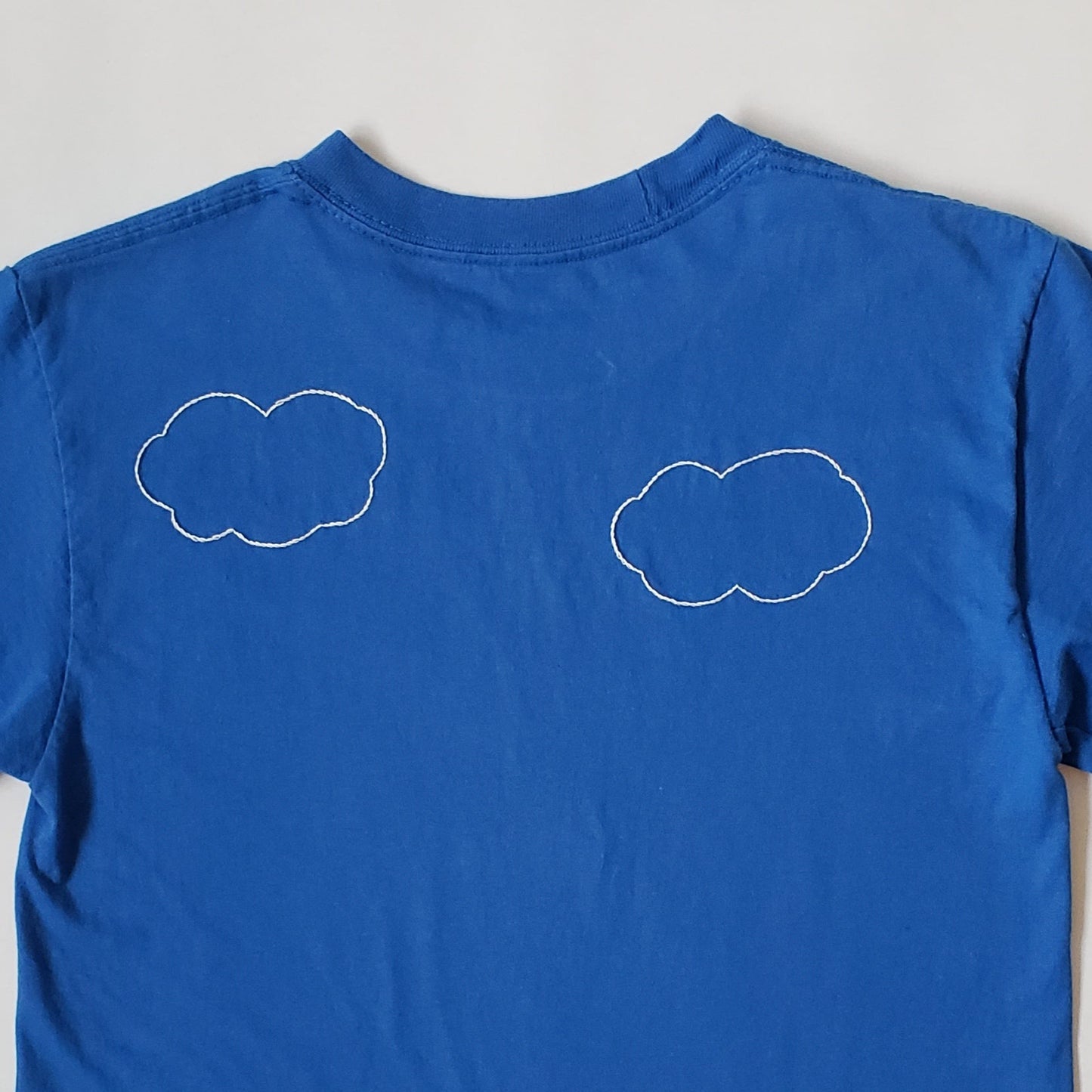 The back of a blue crewneck tee with hand stitched clouds. There are two cutesy white clouds on this shirt. Both very charming and located on the upper back.