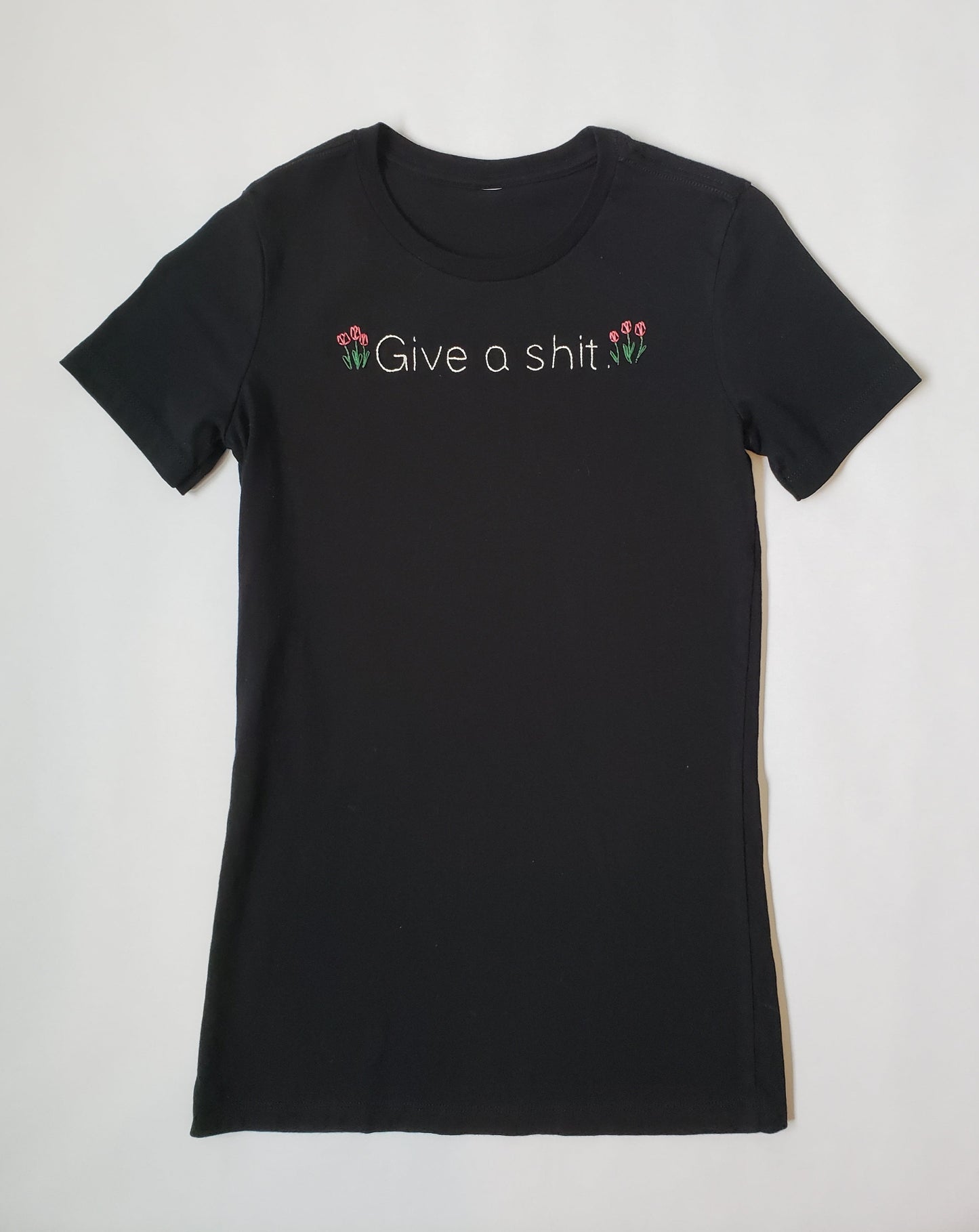 A slim fit black crew neck tee with "Give a shit" hand embroidered in white, in a font similar to Helvetica. Three tulips are on either side of the text. The tulips are gathered together like they are planted alongside the text. They are a muted red.