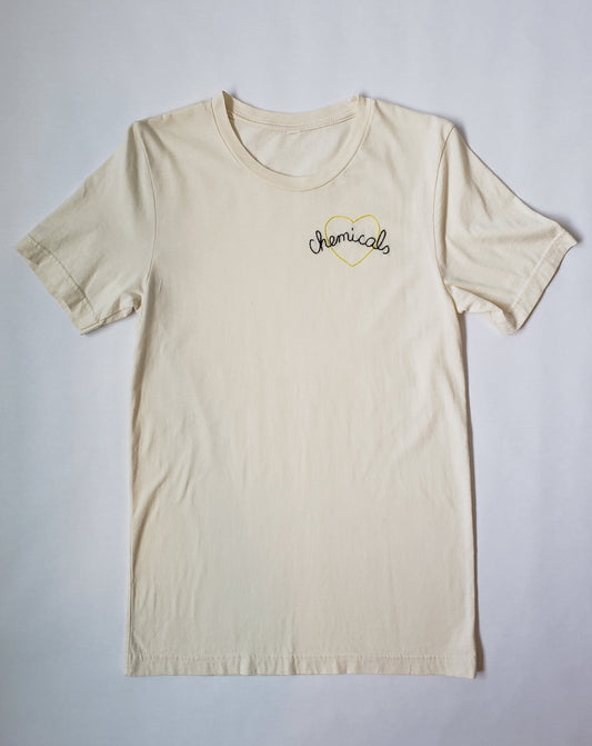 A full shot of a vintage white shade cotton tee with Chemicals stitched in an arc with a yellow heart behind it. Chemicals is stitched in an open split backstitch, meaning it has a chain effect. It is hand stitched in cursive. The heart is done in a vine stitch, so it has a twisted effect.