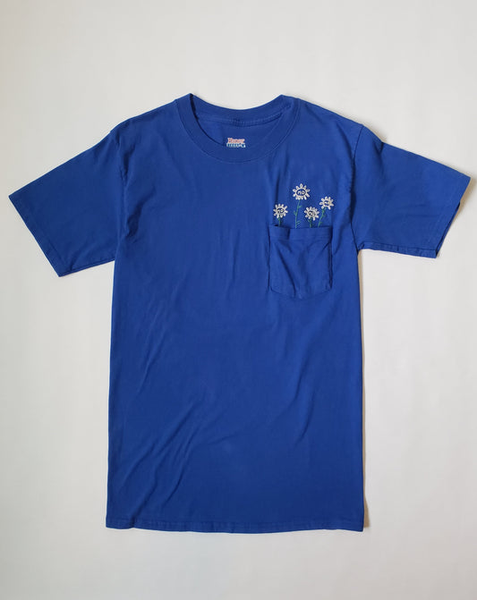 A blue cotton pocket tee is laid flat on a white background. There are four sunflowers popping out of the pocket, all of which have a small lowercase "no" embroidered on the face of the flowers.