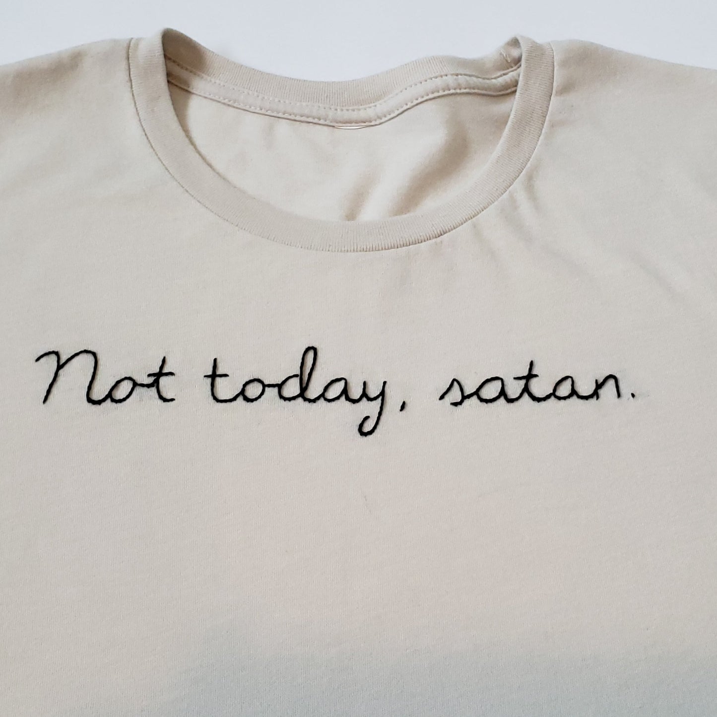A close up of the stitching on a sandstone tee, laid flat on a white background. "Not today, satan." is hand stitched on the tee in black thread in cursive. The artist used a vine stitch.