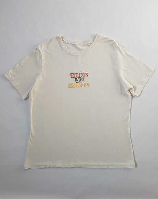 "Take up space" hand embroidered on a cream tee laid on a white background. The font is a bit groovy, flared out at the top and ends of letters, and the palette is decidedly 70s. "Take" is done in a rusty orange, "up" is stitched in a cool brown, and "space" is stitched in a golden yellow shade. All done using whipped backstitch, which gives a twisted effect.