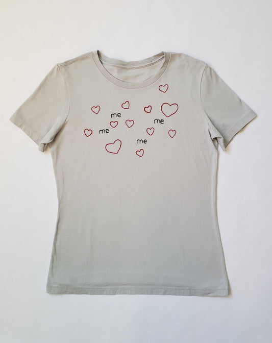 A light gray shirt lays flat on a white background. The shirt has 4 hand embroidered "me" and 12 hearts, two of which are about double the other's size. The text is black and in a typed looking font, and the hearts are dark red and quite round and cute.