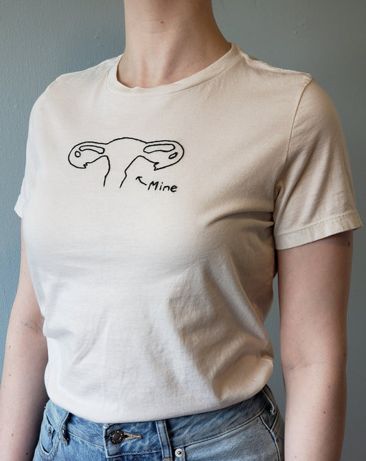 A woman wears a crewneck cream cotton shirt with an anatomically accurate outline of ovaries hand embroidered in black, with "mine" and a small arrow pointing at the outline. The T shirt is a relaxed fit.