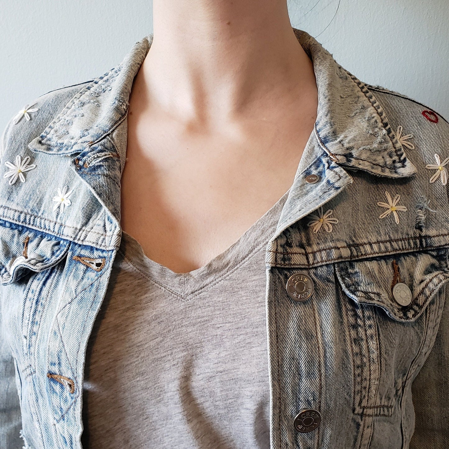 A close up shot of the jean jacket, worn by a woman. We can see that there are at least 4 daisies visible on each shoulder when the collar is down. The daisies are about an inch across and have about 6 petals each. The jacket is worn open and relaxed.