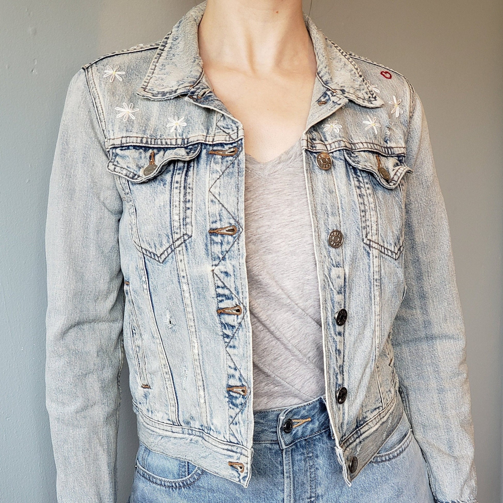 A woman wears an open acid wash denim jacket with daisies covering the upper section of the jacket. A small heart is on the left shoulder. The jacket ends at her upper hip.