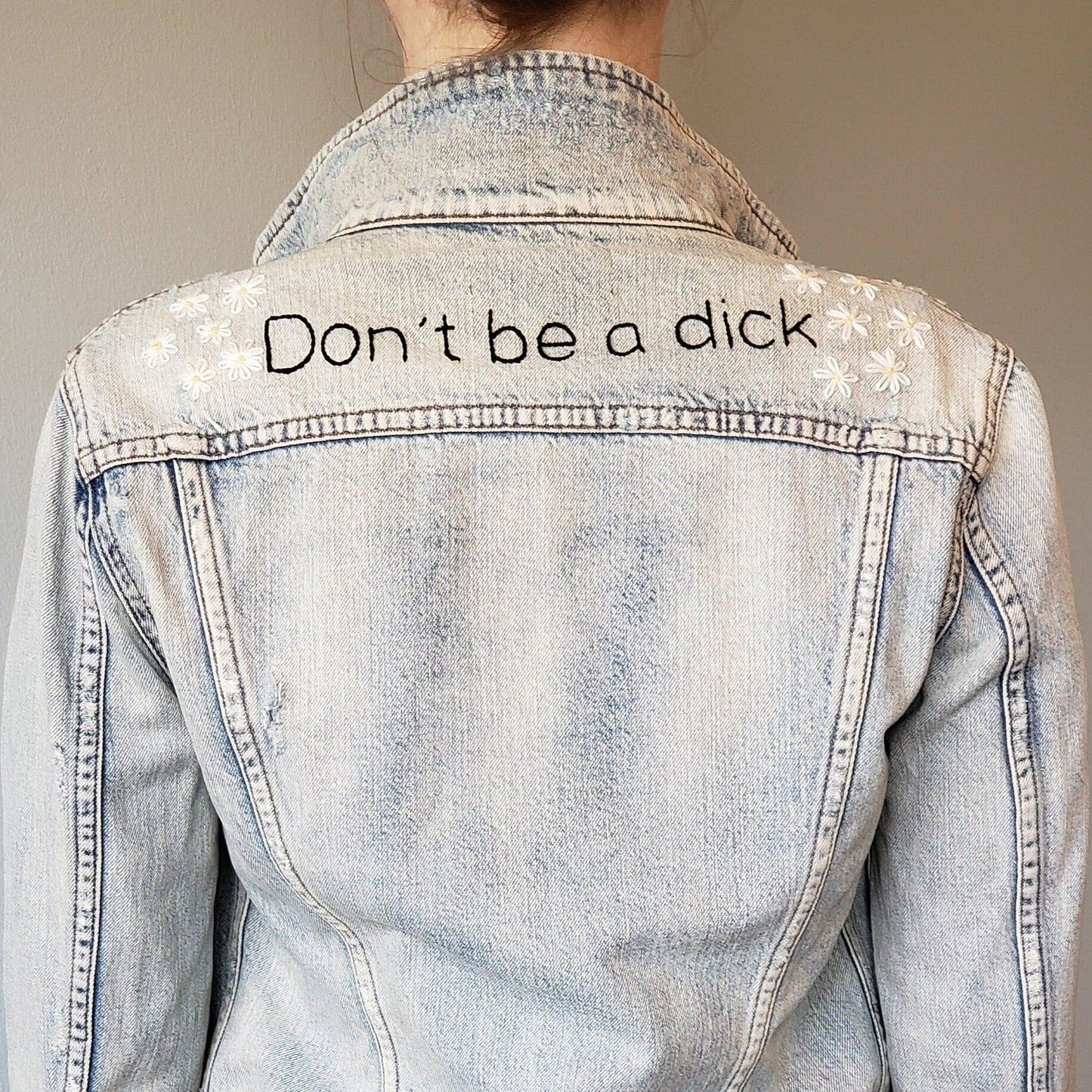 A white woman wears an acid wash denim jacket in front of a light grey background. The jacket has "Don't be a dick" hand embroidered in black, and daises sewn on both shoulders. There are 6 daisies on each shoulder.