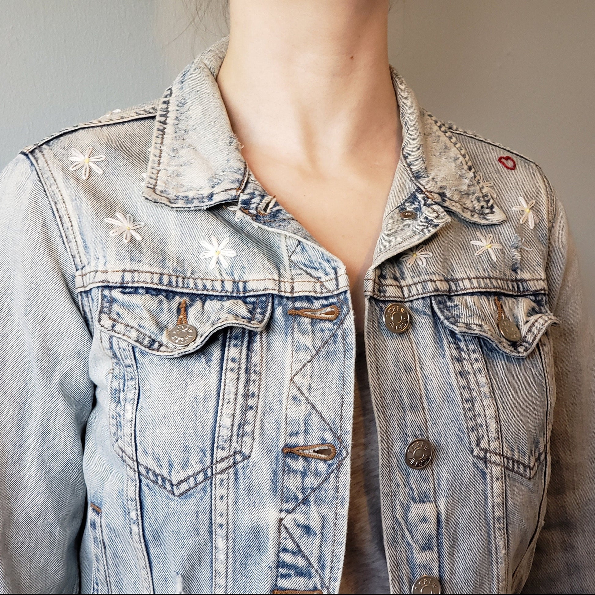 We see the jacket from the right now. The daisies are hand embroidered in an arc on the right chest section. The jacket is a button up acid wash jean jacket with buttoned chest pockets and classic detailing.