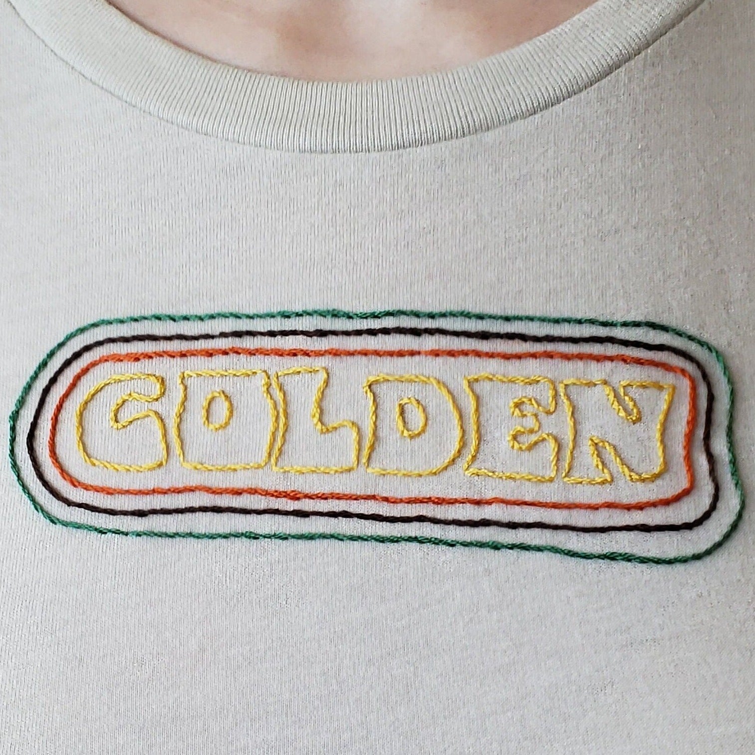 A close up of the text on a sandstone crewneck tee. "GOLDEN" is hand sewn in a groovy font, and the text is encircled by three hand sewn lines. The closest is orange, followed by brown and then green. The decoration sits about 2.5 inches down from the collar.