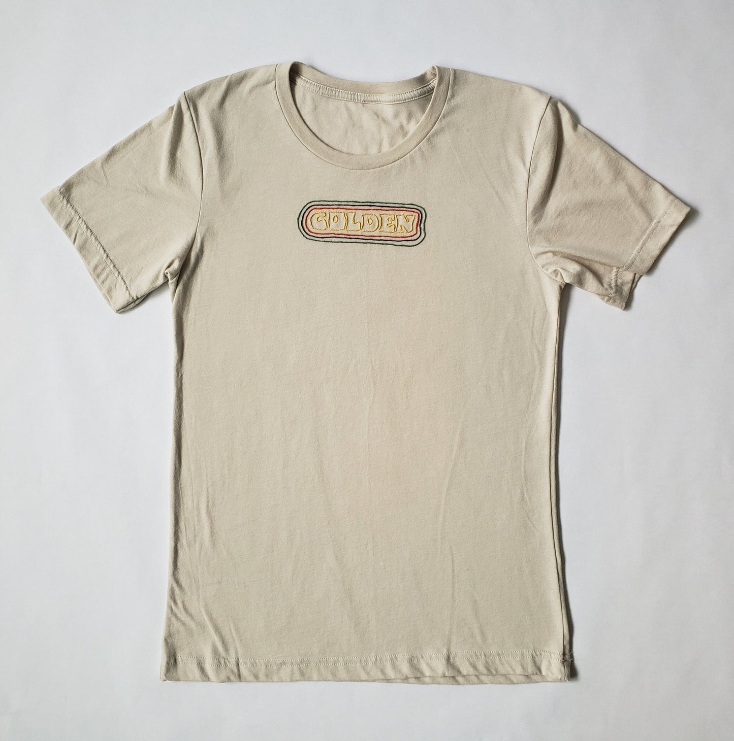 A sandstone crewneck tee lays flat on a white background. "GOLDEN" is hand sewn in a burnished gold thread. The text is encircled by an orange line, then brown, then green. About 4 mm is between each concentric border.