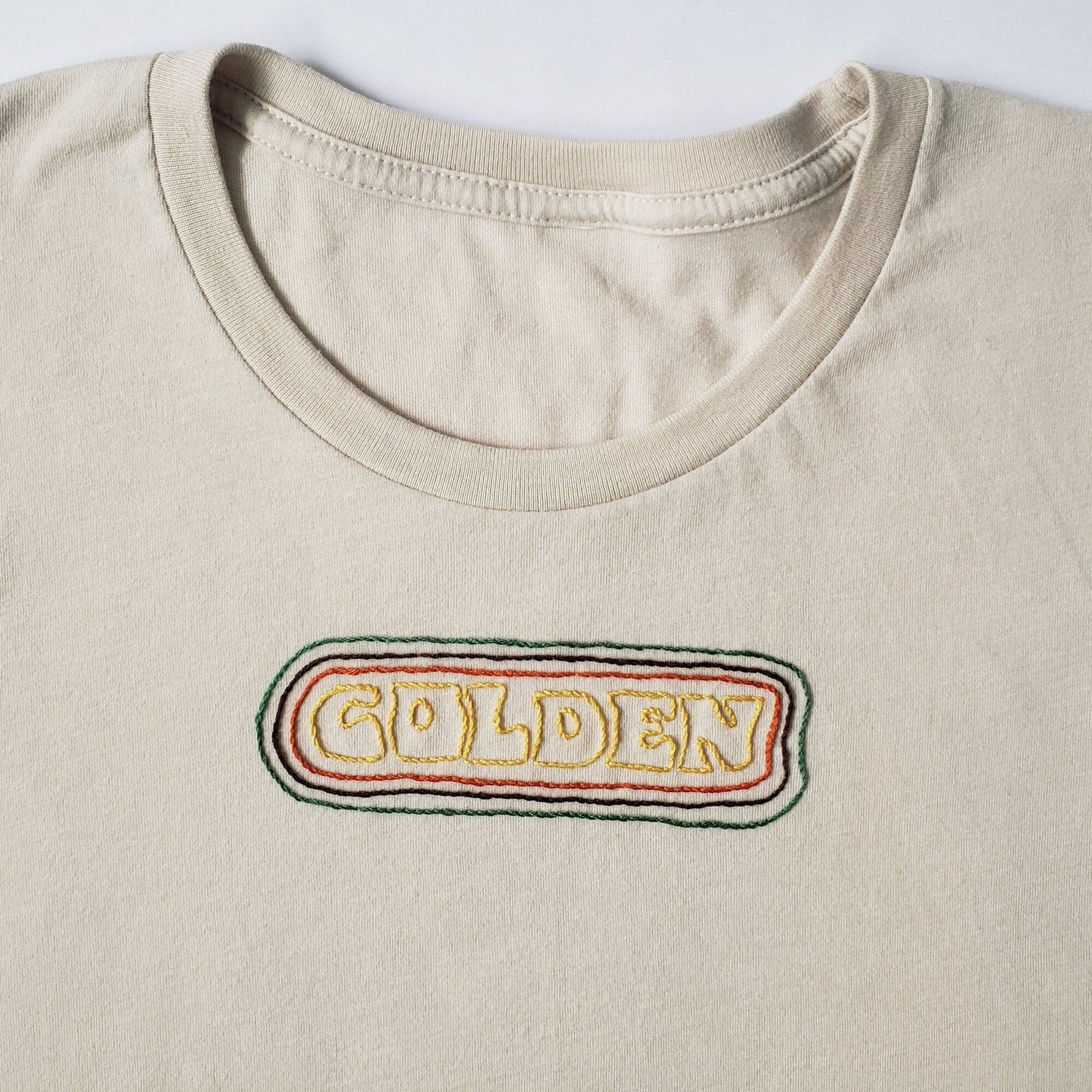 A close up of a soft looking sandstone crewneck tee. "GOLDEN" is hand sewn in a groovy font in burnished gold thread. The text is encircled by an orange line, then brown, then green. About 4 mm is between each concentric border. The effect is retro and awfully fun, I think.