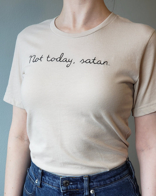A woman wears a light sandstone shade crewneck tee tucked into mid blue denim jeans. The photo is taken from mid neck to upper hip. The background is grey. The shirt says "Not today, satan." and is hand stitched in black thread in cursive.