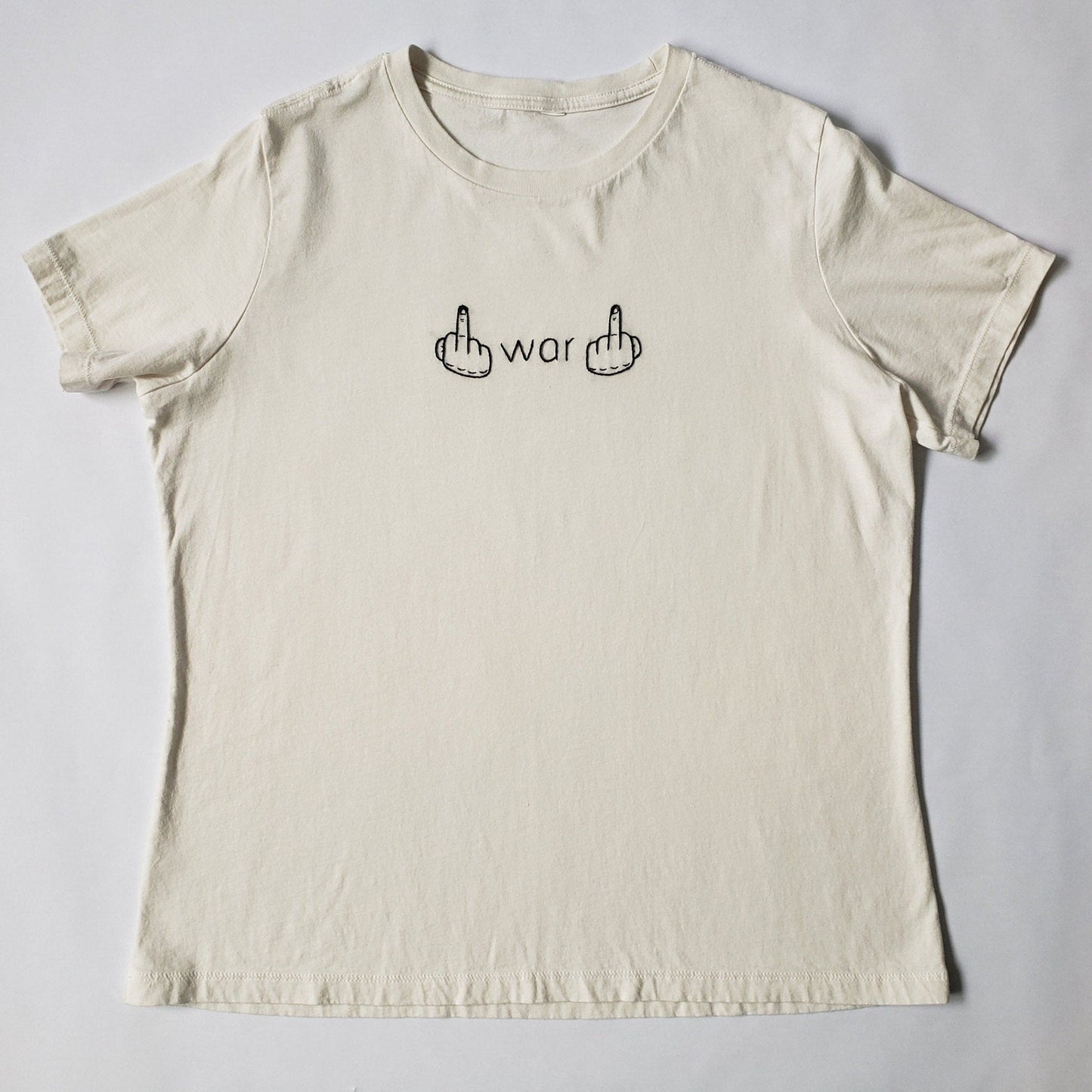 An off white crew neck t shirt lays flat on a white background. War and two hands giving the finger are hand embroidered in the middle of the chest. The text is in a font similar to Helvetica and the hands are slightly brutalist in style