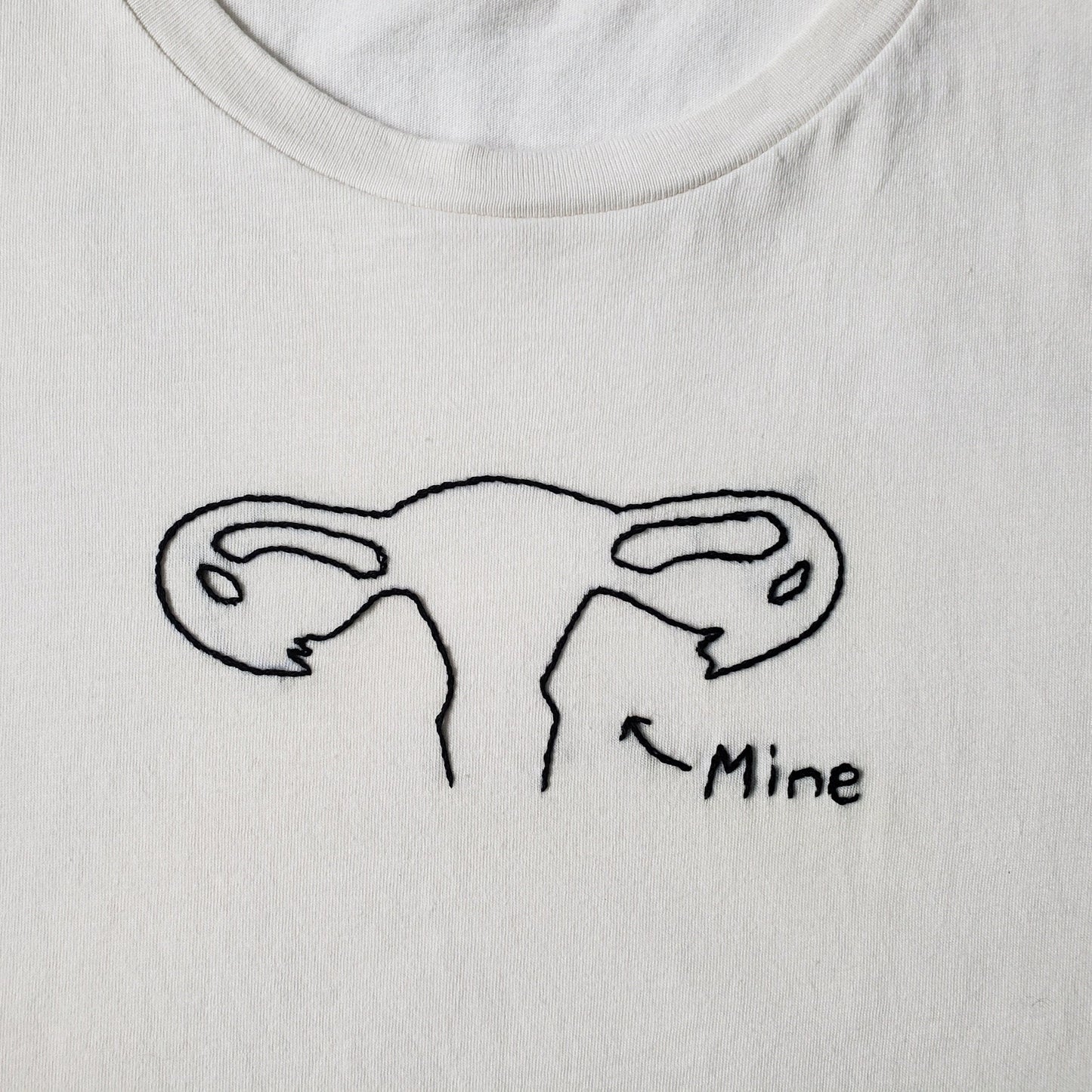 A close up of a hand embroidered uterus on a cream cotton crewneck tee. The silhouette of the organ includes the infundibulum, fimbriae and a bit of the vagina, etc. No external organs, just a fairly complete female internal anatomy in black thread, with a small "Mine" and a little arrow off to the bottom right.