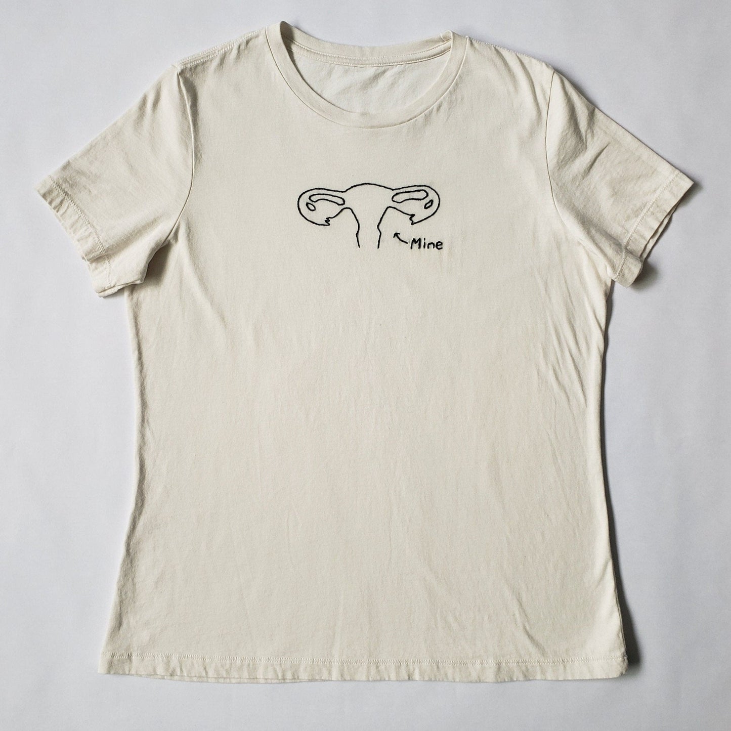 A crewneck cream tee laid flat on a white background. The shirt has a uterus hand embroidered in a vine stitch, and "mine" and a small arrow embroidered in a backstitch. All art is done in black. It is all quite minimal, just the outline of the organ.