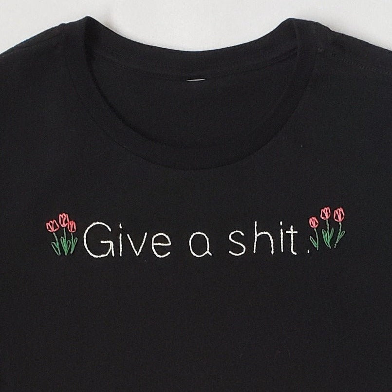 A close up of a black crewneck tee with "Give a shit" hand embroidered in a Helvetica like font. Three tulips sit on each side of the text. The tulips are a muted deep red, and each has two leaves.