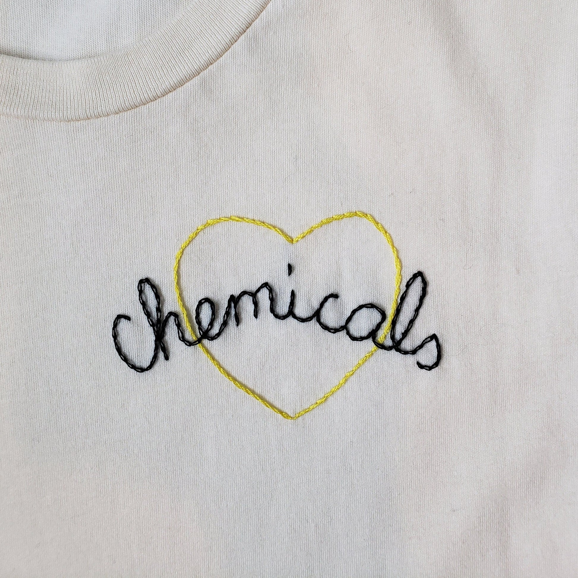 A close up shot of the detail on the left of the vintage white tee. Chemicals is hand stitched in cursive, in an open split backstitch. It is black. It looks like links in a chain. The yellow heart behind the text is done in a vine stitch, so it looks twisted.