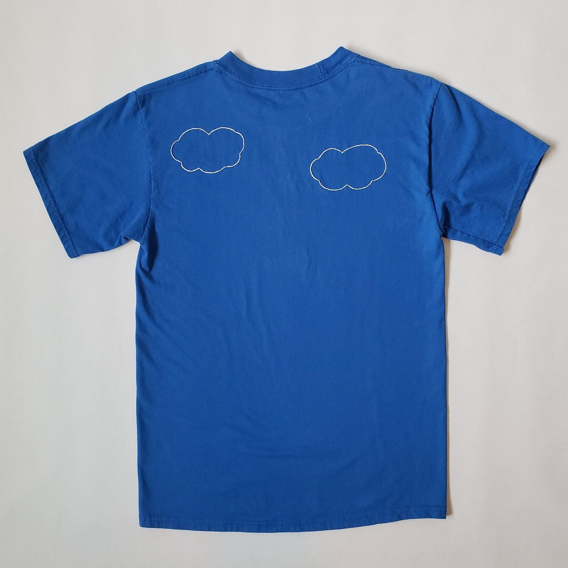 The back of the crewneck blue tee. This side has two clouds, and the one on the left is slightly higher. The clouds are hand stitched in white and have a cute cartoonish look to them.