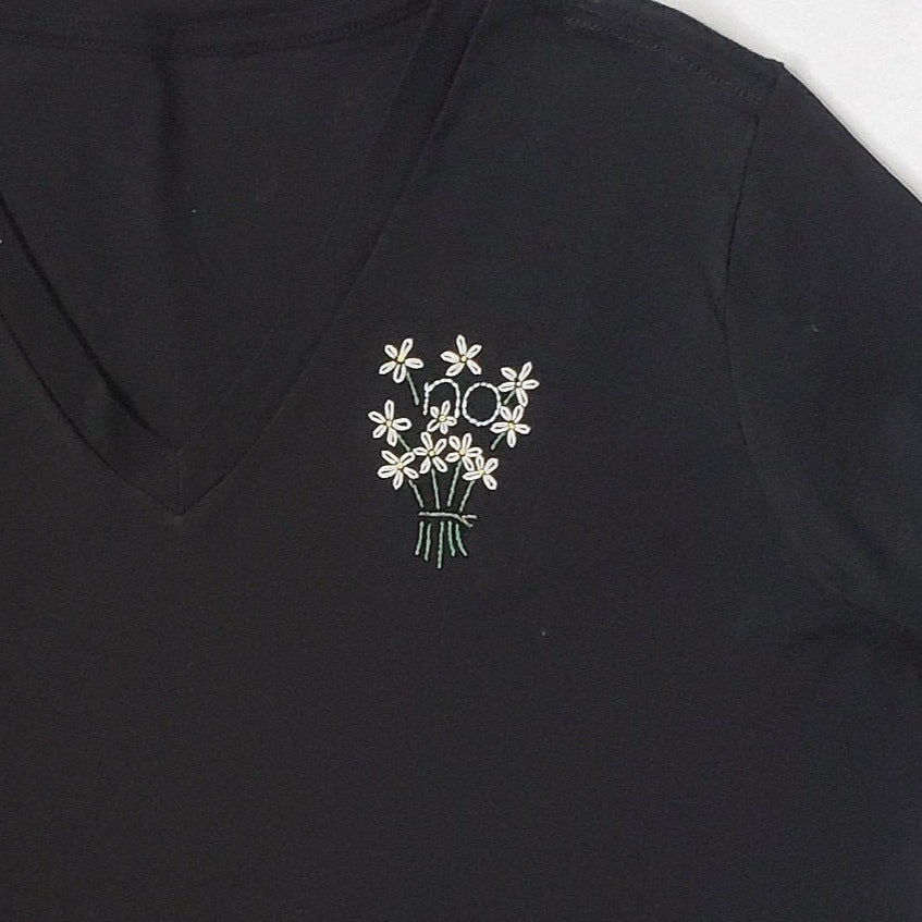 A close up of a hand embroidered daisy bouquet on a black tee. A "no" is embroidered in the middle of the bouquet. The bouquet fastener is grey and the stems have a visible break under the tie, to create visual cohesiveness with the top.