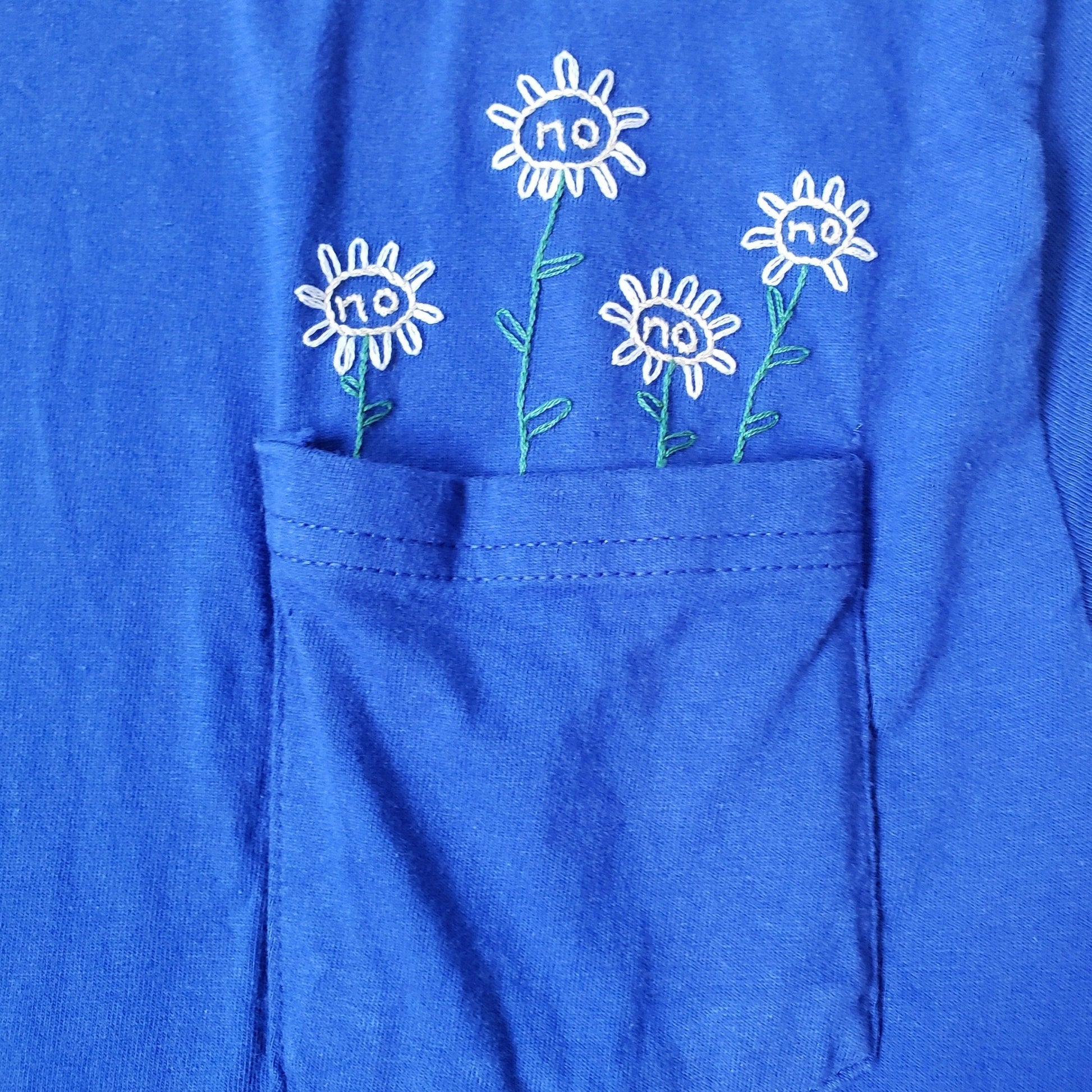 A close up of the pocketed blue tee. Four sunflowers with "no" embroidered in the face of the flowers. The flower furthest to the left is short, the one to its right is tall, the next short, the following taller. There is spacing between each petal.